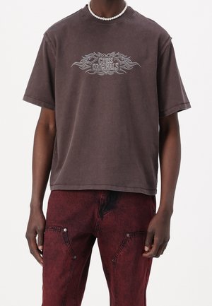 T-shirt Guess Originals marron foncé avec un dessin de flammes, porté avec un pantalon rouge délavé à l’acide et un collier de perles blanc sur une personne.