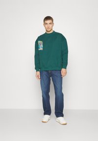 Groene sweatshirt met een grafische zak op de linkerkant van de borst, gecombineerd met blauwe jeans en witte sneakers. Casual pasvorm met ribbels aan de manchetten en onderkant.