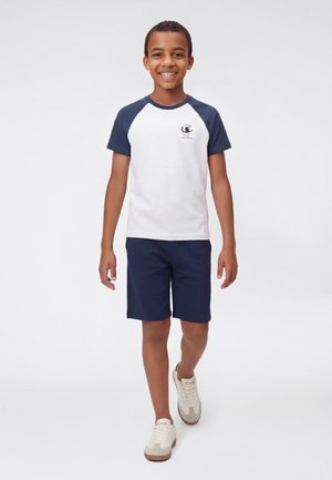 Smiling boy walking forward wearing a white and navy raglan sleeve t-shirt, navy shorts, and white sneakers on a plain white background.