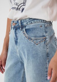 Lyseblå denimjeans med høj talje, der har kobbernitter langs forlommerne og et klassisk design med fem lommer.