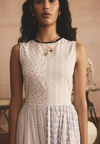 Bela obleka s cvetličnimi in črtastimi vzorci; oprijeta zgornji del, ki prehaja v nabran, razširjen krilo. Zlati nakit okrog vratu.