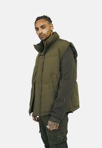 Gilet bouffant vert olive avec un col montant, doté d'une fermeture éclair à l'avant et de poches latérales, superposé à une chemise à manches longues vert foncé.