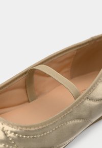 Ballerines en satin beige avec un dessus texturé, une sangle élastique et une doublure lisse, présentant des coutures le long du bord.