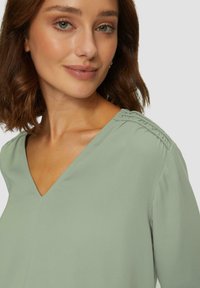 Blouse à col en V en tissu vert doux et atténué avec des détails plissés aux épaules ; la texture lisse complète le design décontracté.