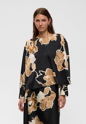 Femme aux longs cheveux bruns portant un haut ample noir et une jupe avec de grands motifs floraux beige et blanc, debout devant un fond uni.