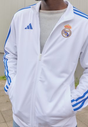 Person trägt eine weiße Real Madrid Jacke mit blauen Adidas-Streifen und Logos, Hände in den Taschen, steht draußen auf einer gepflasterten Fläche.