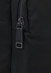 Dettaglio della cerniera di una borsa nera con superficie testurizzata, dotata di un tirante della cerniera nero opaco etichettato "BOSS" e di elementi di design eleganti e minimalisti.