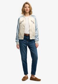 Giacca bomber leggera in raso con maniche blu, corpo beige e uccelli ricamati. Abbinata a jeans blu e scarpe slip-on color cuoio.