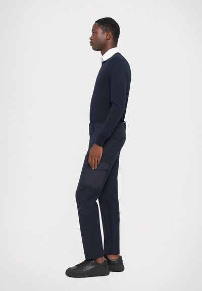 Pulover bleumarin și pantaloni ajustați asortați, cu buzunar lateral tip cargo. Material tricotat texturat și țesătură netedă, asortate cu teniși negri cu talpă înaltă.