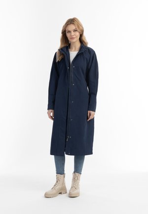 Marineblauwe lange jas met een ritssluiting aan de voorkant, drukknopen en een capuchon. Het model draagt lichtblauwe jeans en beige veterlaarzen. Gladde textuur.