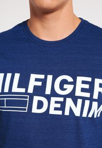 Modré bavlněné tričko s kulatým výstřihem, které nese výrazný bílý text "HILFIGER DENIM" a jednoduchý grafický prvek.