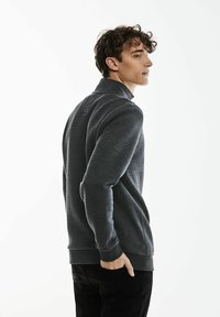 Grå, texturerad tröja med hög krage, med ribbade mudd och nederkant. Modellen står i en lätt vinkel mot en plain bakgrund.