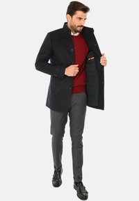 Cappotto in misto lana blu navy con colletto alto, chiusura a bottoni monopetto e foderato in tessuto liscio. Indossato sopra un maglione bordeaux e pantaloni grigi.