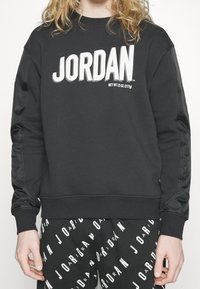 Personne portant un sweat-shirt noir avec le texte « JORDAN » et un pantalon noir orné de motifs répétant le logo « JORDAN » en blanc.