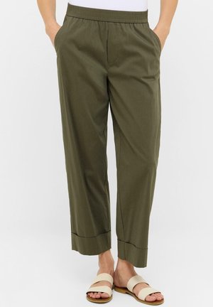 Osoba w oliwkowozielonych spodniach z elastycznym pasem i mankietami oraz beżowych klapkach, stojąca z rękami w kieszeniach.