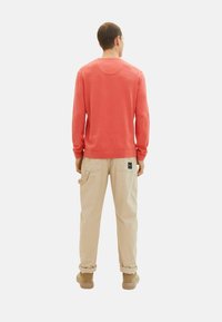Koralfarvet sweater med lange ærmer, rund hals og manchetter i bunden, parret med lysebeige cargo-bukser med lommer og mærke. Brune sko fuldender outfittet.