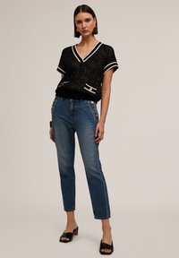 Zwarte gebreide trui met een V-hals en korte mouwen, met crèmekleurige accenten. Hoge blauwe jeans met knoopdetails aan de zijkant. Zwarte hakken.