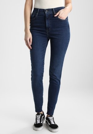 Jean slim bleu foncé porté avec des baskets noires et blanches et un top blanc côtelé, main dans la poche, debout sur un fond blanc.