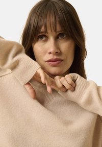 Beige Strickpullover mit V-Ausschnitt, gerippten Bündchen und weicher Textur. Das Modell passt den Kragen an und zeigt den Fall und die Verarbeitung des Stoffes.