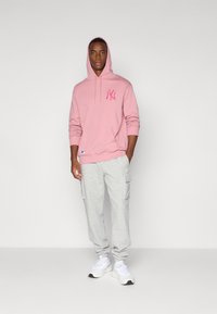 Sweatshirt à capuche rose clair avec poche avant et logo rose, associé à un pantalon cargo gris avec poches latérales et ourlets, porté avec des baskets blanches.