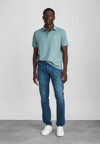 Απαλή μπλε πόλο μπλουζίτσα με κοντά μανίκια και διακριτό λογότυπο, σε συνδυασμό με τζιν σε σκούρο μπλε απόχρωση και λευκά αθλητικά παπούτσια. Απλός σχεδιασμός, casual εφαρμογή.