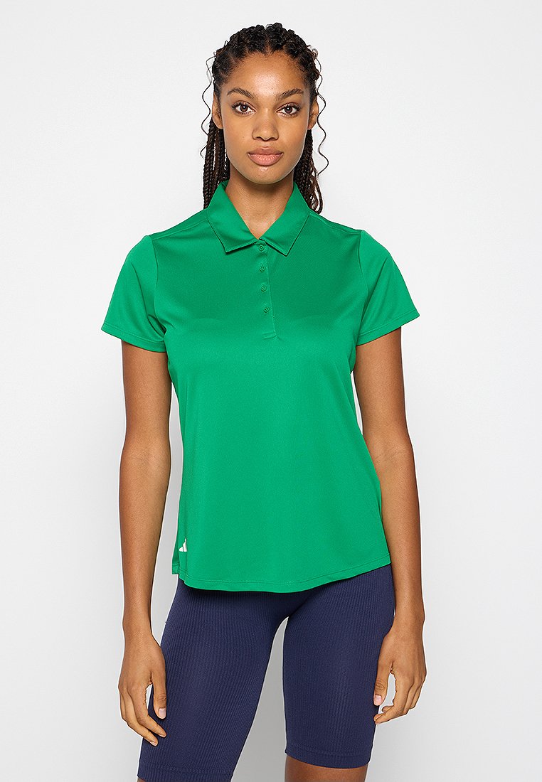 Femme aux cheveux tressés portant un polo à manches courtes vert vif et un short de cyclisme bleu marine, debout devant un fond blanc.