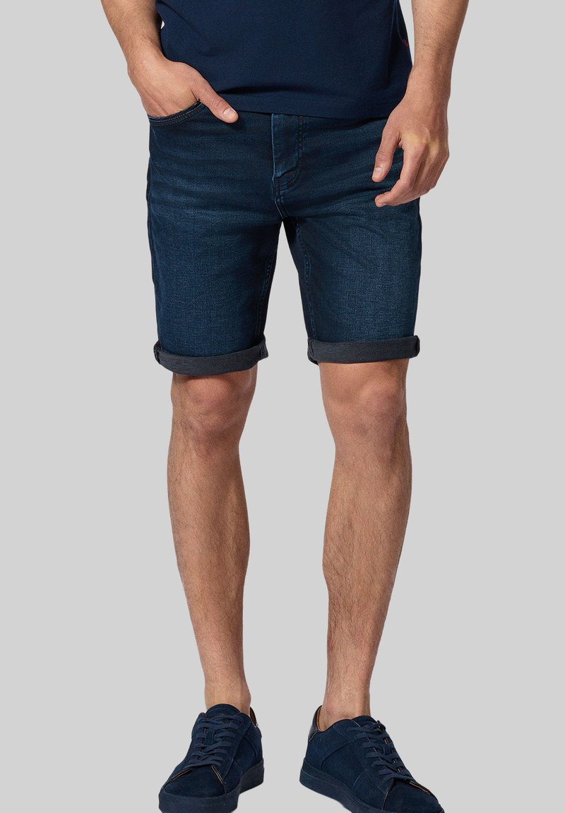 Ciemne szorty dżinsowe z podwiniętym dołem, prosty krój i pięć kieszeni. Noszone z granatową koszulą i granatowymi trampkami. Gładka tekstura.