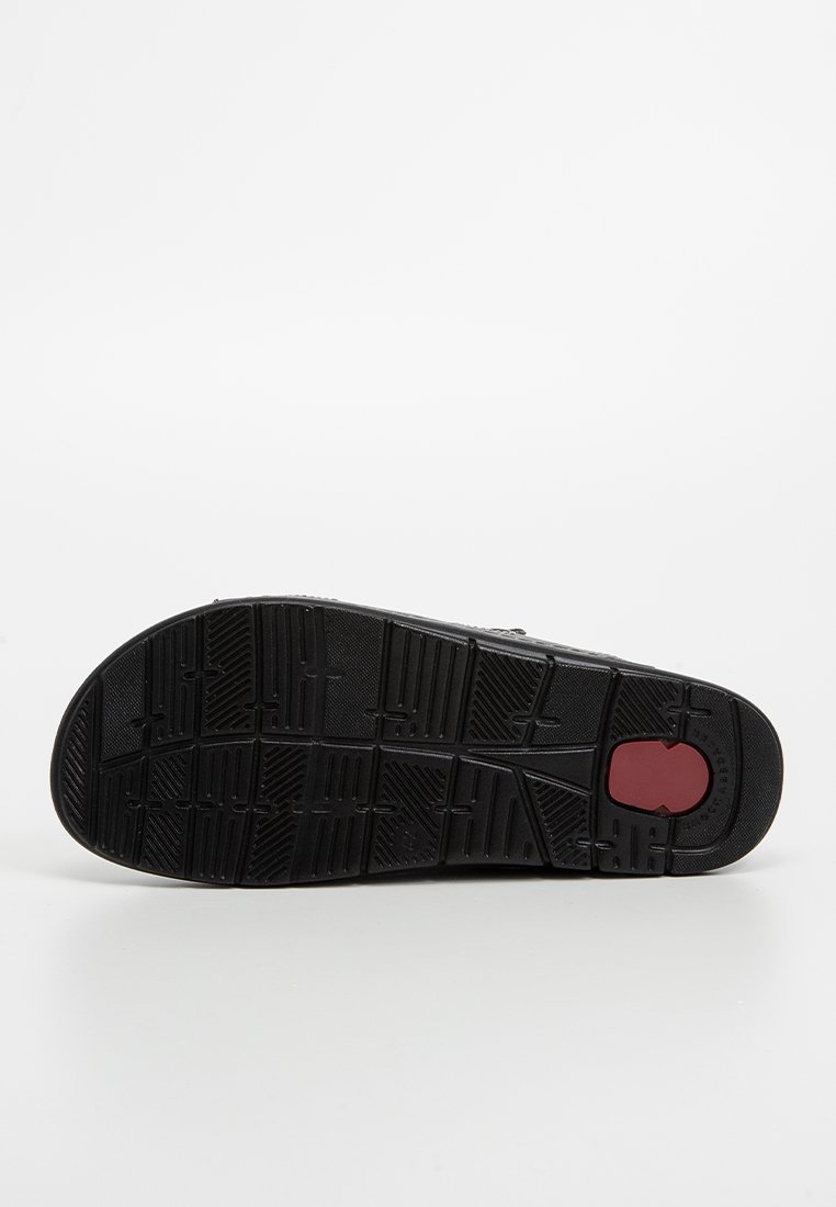 Semelle de chaussure noire avec un motif de bande de roulement texturé et un insert circulaire rouge près du talon, affichée sur un fond blanc uni.