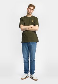 Camiseta de algodón verde oliva de manga corta con un logo en el pecho, combinada con jeans de mezclilla azul y zapatillas crema con acentos en navy.