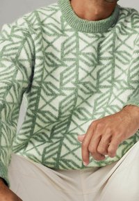 Pull en tricot présentant un motif géométrique vert et blanc, avec une finition texturée et un col côtelé. Main reposant sur l'ourlet inférieur.