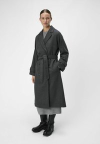 Manteau en laine gris avec boutons double boutonnage, taille ceinturée et manches retroussées, associé à un pull gris en tricot et des bottes noires.