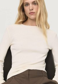 Rippendes Langarmoberteil in Creme, mit einem Bootsausschnitt und figurbetonter Silhouette. Getragen mit dunkelbraunen Hosen, sitzend vor einem neutralen Hintergrund.