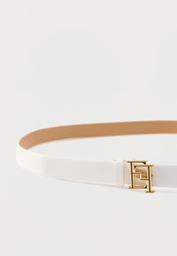 Λευκή συνθετική ζώνη με λεία υφή, που διαθέτει αγκράφα από χρυσό μέταλλο με γεωμετρικό σχέδιο και ταμπά εσωτερική επένδυση.