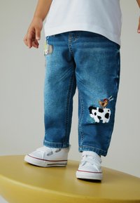 Siniset farkut, joissa on kirjailtuja lehmä- ja tipuaiheisia kuvioita, väljällä istuvuudella ja käännetyillä lahkeensuilla. Yhdistetty valkoisten tennarien kanssa.