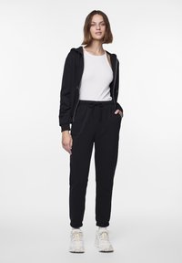 Musta vetoketjullinen huppari taskuilla, jonka alla on valkoinen tanktop ja se on yhdistetty mustiin collegehousuihin. Tennarit ovat valkoiset keltaisilla yksityiskohdilla.