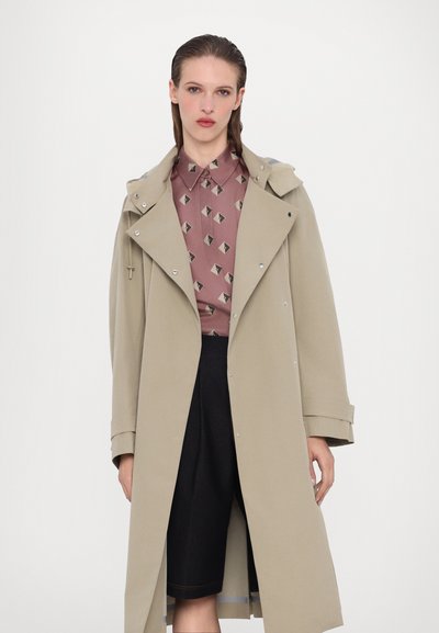 Beżowy trencz z dużymi klapami i srebrnym wykończeniem, noszony na wzorzystą różową koszulę i czarną spódnicę. Gładka faktura i długość do kolan.