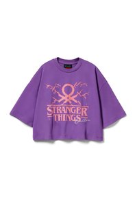 Camiseta morada recortada de algodón con un gráfico audaz en rosa que dice "STRANGER THINGS" con elementos de diseño adicionales. Corte holgado y mangas cortas.
