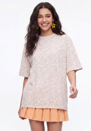 Smilende kvinde med langt mørkt hår iført en lys mønstret oversized skjorte, orange plisseret nederdel og store gule stjerneformede øreringe.