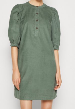 Donna che indossa un vestito verde fino al ginocchio con maniche a sbuffo a metà braccio e una patta frontale con bottoni, in piedi davanti a uno sfondo neutro.