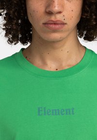 Zelené bavlnené tričko s okrúhlym výstrihom a krátkymi rukávmi, na prednej strane je potlač s nápisom "Element" v tmavomodrej farbe.