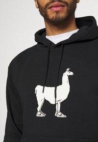 Černá mikina s kapucí, na které je bílá silueta lamy v botách, s šňůrkovou kapucí a klokankou. Měkká textilie.