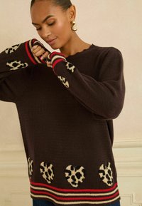 Brązowy sweter dziany z sercowymi wzorami w panterkę, czerwone i beżowe paski na mankietach i dolnej krawędzi. Miękka tekstura, luźny krój, okrągły dekolt.