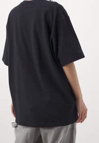 Černé oversized tričko z bavlny s krátkým rukávem, kulatým výstřihem a volným střihem, kombinované se šedými kalhotami. Hladká textura a jednoduchý design.
