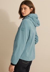 Veste à capuche bleu clair avec fermeture éclair sur le devant, grande poche avant et patch en caoutchouc sur la manche. Texture lisse, coupe décontractée et accents noirs.