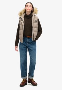 Beige Steppweste mit fellgefütterter Kapuze, brauner Strickpullover, blaue Jeans und braune Schnürstiefel mit gelben Akzenten.