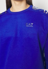 Sweat-shirt bleu à col rond, avec un logo EA7 brillant et un marquage le long de l'épaule en lettres dorées. Texture douce.