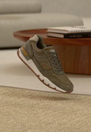 Olivgrüner Turnschuh mit weißer und brauner Sohle, der über einem beigen Teppich schwebt; im Hintergrund ist ein Couchtisch aus Holz und ein Bücherstapel zu sehen.