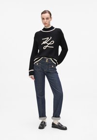 Schwarzer gerippter Strickpullover mit weißem Saum und Logo, kombiniert mit dunkelblauen geraden Jeans, die goldene Knöpfe und umgeschlagene Säume haben.