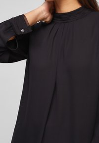 Schwarze Bluse mit langen Ärmeln, hohem Ausschnitt und plissierten Details. Verfügt über dekorative Nähte und einen Knopfakzent am Bündchen.