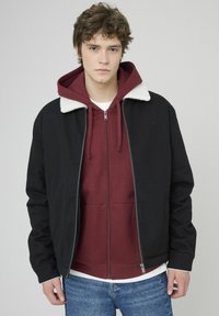 Veste aviateur noire avec col en fausse fourrure blanche sur un sweat à capuche zippé bordeaux. Le modèle porte un jean bleu. Texture lisse, design contemporain.